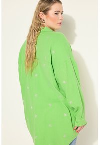 Zelená oversized košile s texturovanou látkou, zdobená stříbrnými hvězdami. Knoflíkové manžety a volný střih.