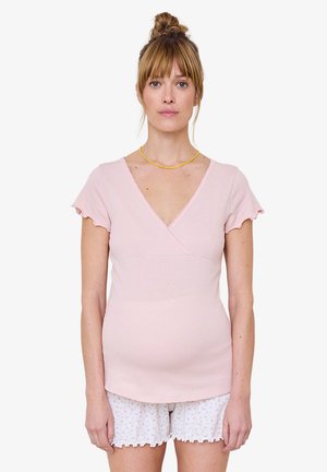 Frau mit zusammengebundenem Haar, die ein hellrosa kurzärmeliges Oberteil und weiße florale Shorts trägt, steht vor einem einfarbigen Hintergrund.