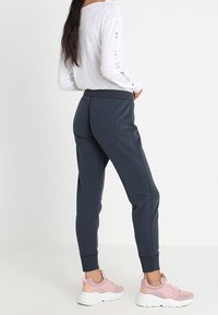 Vrouw in donkerblauwe joggers, wit langwerpig shirt met kleurrijke tekst op de mouwen, en roze-witte sneakers, staat zijwaarts.