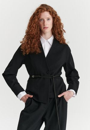 Vrouw met lang krullend rood haar draagt een zwart pak met riem over een wit overhemd met kraag, staand met handen in de zakken tegen een effen achtergrond.