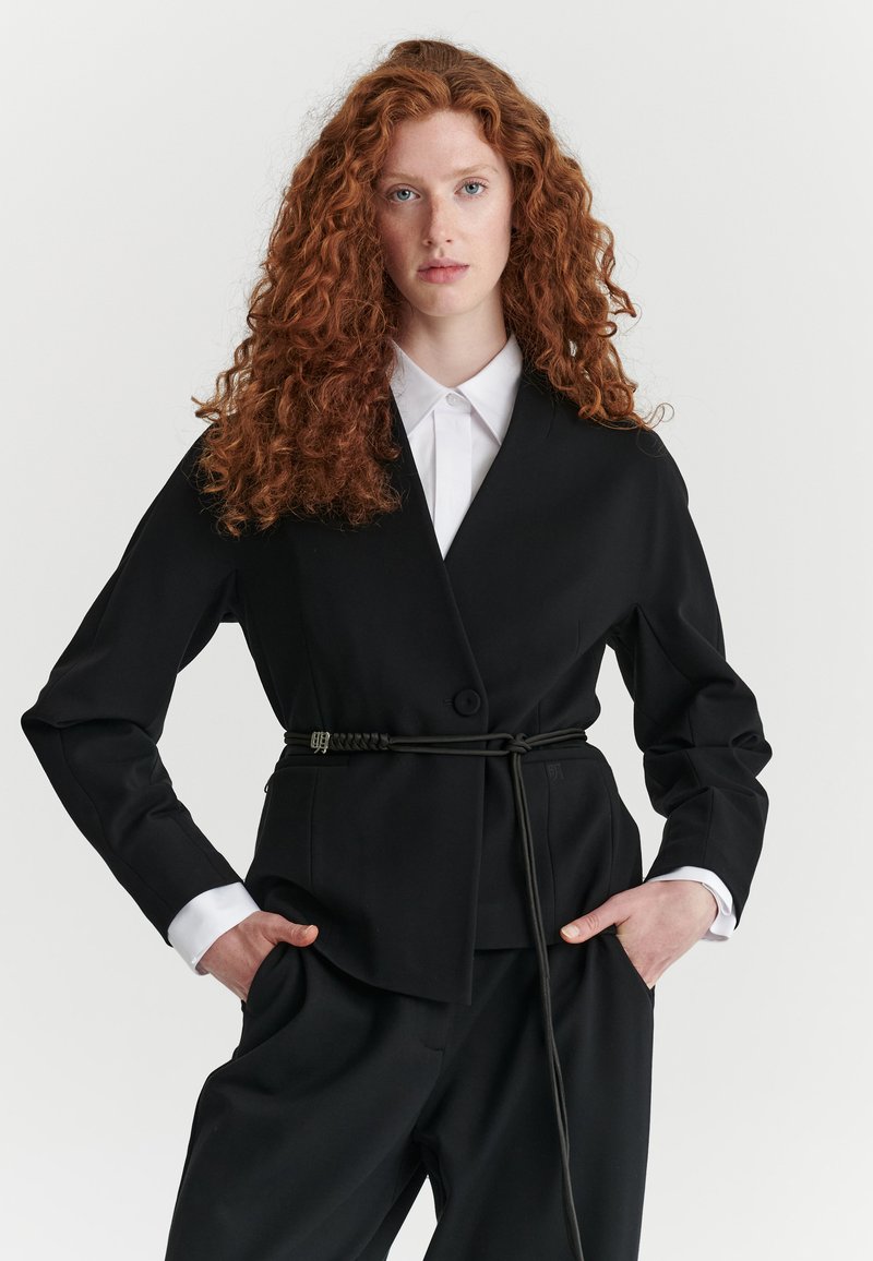Femme aux longs cheveux roux bouclés porte un costume noir avec une ceinture sur une chemise blanche à col, debout les mains dans les poches devant un fond uni.