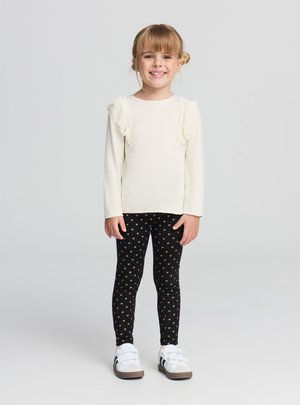 Maglia lunga a maniche lunghe color crema chiaro con spalle arricciate, abbinata a leggings neri con piccoli cuori gialli e scarpe bianche con dettagli neri.