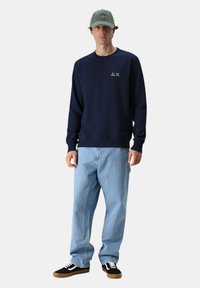 Giovane uomo che indossa un cappellino verde, una felpa blu navy con un piccolo logo bianco, jeans larghi blu chiaro e scarpe nere con lacci bianchi.