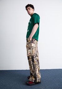 Πράσινη δίχρωμη μπλούζα με λογότυπο, παραλλαγής cargo παντελόνια με τσέπες και σκούρες ρίγες, και μαύρα/κόκκινα αθλητικά παπούτσια με υφή.