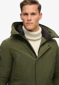 Olivegrüner, gepolsterter Jacke mit Kapuze, mit durchgehendem Reißverschluss, seitlichen Taschen und einem kontrastierenden schwarzen Innenfutter. Getragen über einem cremefarbenen Rollkragenpullover.