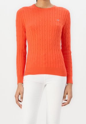 Femme portant un pull torsadé orange vif à manches longues et un pantalon blanc sur un fond clair uni.