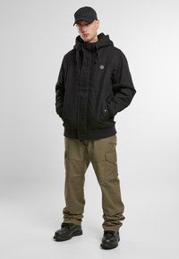 Chaqueta negra con capucha y cierre de cremallera, pantalones cargo beiges con bolsillos laterales y botas negras; modelo usando una gorra negra a juego.