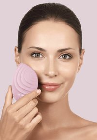 Silikone ansigtsrensningsenhed i blød pink, med bølget overfladestruktur og bløde børster, holdt mod en models kind.