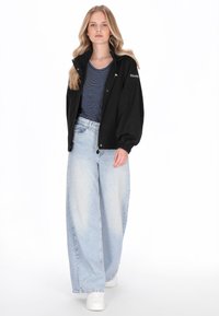 Zwarte jas met ritssluiting en een hoge kraag, gecombineerd met een marineblauw-wit gestreept shirt en lichtblauwe jeans met wijde pijpen. Witte sneakers.