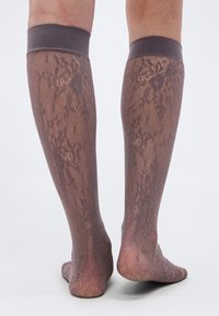 Πόδια με ημιδιάφανα καφέ δαντελένια καλσόν μέχρι το γόνατο με λουλουδένια σχέδια, που φαίνονται από πίσω με φόντο απλό.