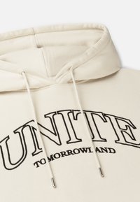 Crème kleurige hoodie met een grote zwarte opdruk met de tekst "UNITE" en "TOMORROWLAND." Heeft trekkoorden en een zachte, textuurachtige stof.