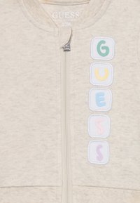 Bio-Baumwolle Kapuzenanzug in hellbeige mit verspielten, mehrfarbigen quadratischen Patches, die die Buchstaben "G", "U", "E", "S", "S" zeigen.