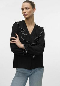 Sort bluse med V-udskæring, der har hvide rillet detaljer langs fronten og ærmerne. Stoffet ser let og luftigt ud.