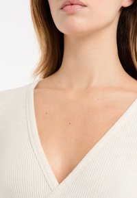 Jasnobeżowy prążkowany top z głębokim dekoltem w kształcie litery V, odsłaniający gładką tonację skóry i minimalne detale tekstury wokół linii szyi.