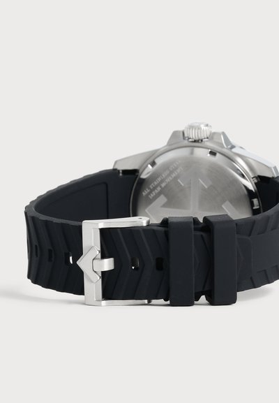 Gros plan sur le fond de boîtier argenté d’une montre-bracelet et son bracelet en caoutchouc noir strié, avec une boucle argentée et deux passants.