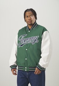 Jaqueta varsity verde e branca com "Tommy" bordado em grande estilo, apresentando botões de pressão, punhos canelados e um corte descontraído.
