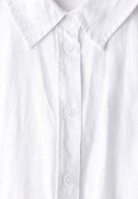 Camicia bianca in lino con colletto classico, sei bottoni sul davanti e un tessuto leggermente strutturato per una sensazione traspirante.