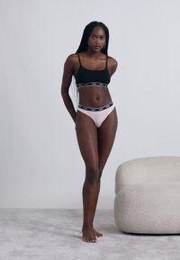 Zwarte beha met brede bandjes en logoband, gecombineerd met lichtroze bikinibroekjes met een vergelijkbare logoband, geplaatst tegen een minimalistische achtergrond.