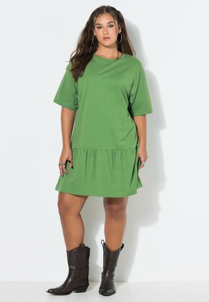 Junge Frau mit langen Haaren, die ein lockeres grünes Kurzarmkleid und dunkelbraune Stiefel bis zur Mitte der Wade trägt, steht vor einem schlichten weißen Hintergrund.