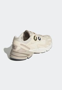 Beige träningssko med ett texturerat mesh-överkapa, lagerdesign och ögla i guld och marinblått. Dämpat sula med ljusgrå accenter.