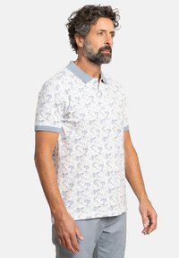 Camisa polo blanca de manga corta con un patrón floral azul, cuello y puños contrastantes en azul claro, hecha de tela transpirable.