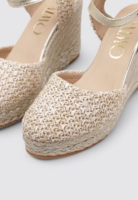 Sandalias de cuña con una parte superior de tejido color beige, acentos metálicos dorados y una plataforma de cuerda texturizada. Correa ajustable en el tobillo para un ajuste seguro.