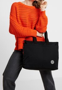 Sac fourre-tout noir à texture lisse, avec une fermeture éclair sur le dessus et deux poignées. Caractérisé par un logo circulaire. Porte avec un pull en maille orange et un jean foncé.