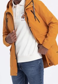 Veste imperméable jaune avec capuche, poches zippées et accents gris, portée sur un sweat-shirt blanc et un jean bleu.