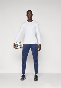 Camisa branca de mangas longas, calças de desporto azul marinho, ténis pretos, segurando uma bola de futebol preto e branco com detalhes de logotipo. Fundo simples.