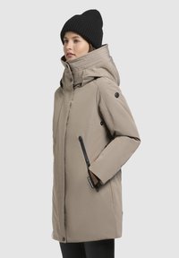 Beige'isolatsiooniga väliskate, millel on suur kapuuts, ees lukuga, küljetaskud ja mustad aktsendid. Kantud musta kudumi beanie'ga.