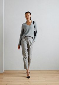Un pull gris côtelé associé à un pantalon tailleur gris clair et des talons noirs. Un petit sac à main noir pend sur le côté. Fond sparse.