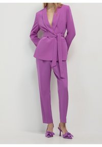 Fioletowy garnitur z dwurzędowym żakietem, który ma pasek w talii oraz dopasowane, slim fit spodnie. Całość uzupełniają czółenka z kokardkami.