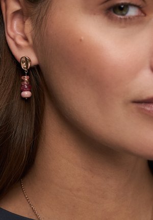 Gouden studs met drie gestapelde roze en paarse kralen, gedragen door een persoon met lichtbruin haar en een gladde huid.