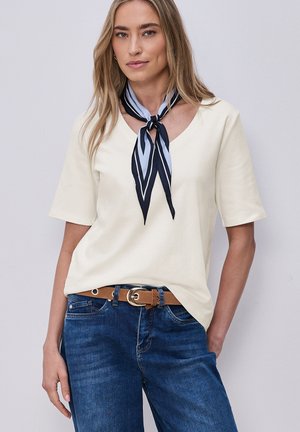 Kobieta o długich blond włosach, ubrana w kremowy top z krótkim rękawem, niebieskie dżinsy z brązowym paskiem oraz czarno-biały paskowany szalik przy szyi, na szarym tle.