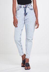 Jean taille haute bleu clair en denim avec un délavage, doté de poches avant et de détails de coutures visibles, associé à des sandales noires.