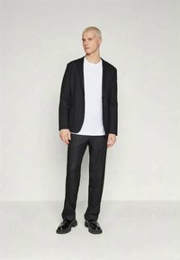 Sort jakkesæt med en glat tekstur, hvid t-shirt under, sorte bukser og blanke sorte sko. Model står med en neutral udtryk.