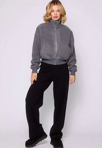 Giacca grigia con cerniera in tessuto strutturato, con collo alto e fascia elastica in vita, abbinata a pantaloni neri a gamba larga e scarpe nere.