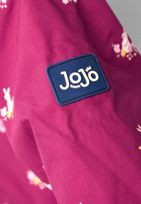 Violetinė medžiaga su gėlių raštais ir trumpikėmis, o mėlynoje stačiakampėje etiketėje įspausta "JoJo" balta rašyba.