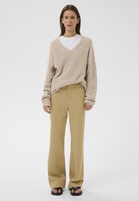 Jeune femme debout portant un pull en tricot beige oversize, une chemise blanche, un pantalon ample couleur beige clair et des sandales noires sur un fond clair uni.