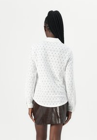Camicia bianca a maniche lunghe con un intricato motivo di pizzo; colletto e polsini con bottoni. Abbinata a una mini gonna marrone lucida con texture a croc.