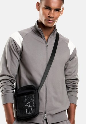 Hombre con una chaqueta gris con cremallera y detalles blancos en los hombros, llevando un bolso negro cruzado con el texto "EA7" en el bolsillo frontal de malla.