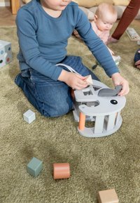 Jong kind in blauw shirt steekt blokken in een olifantvormige blokkendoos, baby en volwassene in de buurt op een groen tapijt.