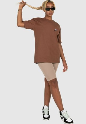 Braunes Oversized-T-Shirt mit einem kleinen Berglogo, kombiniert mit beigen Biker-Shorts und schwarzen High-Top-Sneakers mit weißen Akzenten.
