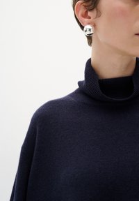 Jūras spēku zilas vilnas kaklasaites džemperis ar ribotu apkakli, valkāts ar apaļu sudraba auskaru, kas atspīd gaismu. Tuva aina ar kaklu un ausi.