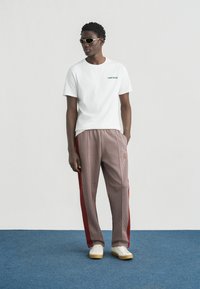 Άσπρο βαμβακερό μπλουζάκι με πράσινο λογότυπο "CONVERSE". Καρό παντελόνια με κόκκινες ρίγες στο πλάι. Το μοντέλο φοράει χρυσά γυαλιά ηλίου και λευκά παπούτσια με κίτρινες σόλες.