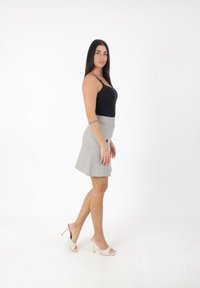 Kobieta z długimi, ciemnymi włosami stoi bokiem, ubrana w czarny top na ramiączkach, szary spódnicę, białe sandały na obcasie oraz tatuaż na lewym przedramieniu.