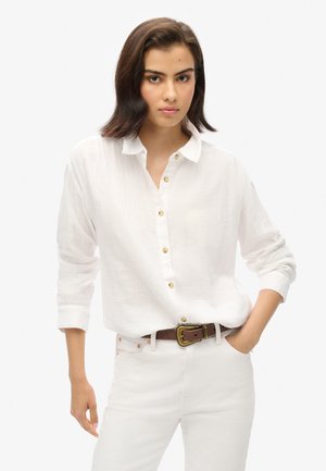 Chemise blanche à boutons, à manches longues, en tissu léger. Dotée de boutons dorés et d'une coupe décontractée. Associée à un pantalon clair et une ceinture marron.