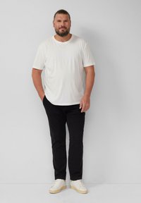 Weißes Baumwoll-T-Shirt, schwarze Hose und weiße Sneakers mit beiger Sohle. Lässiger Schnitt, steht vor einem einfarbigen, hellen Hintergrund.