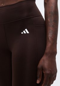 Κοντινό πλάνο ενός ατόμου που φοράει σκουρόχρωμο καφέ κολάν με ένα μικρό λευκό λογότυπο Adidas κοντά στο ισχίο και ένα τατουάζ στο χέρι που ακουμπάει δίπλα στον μηρό.