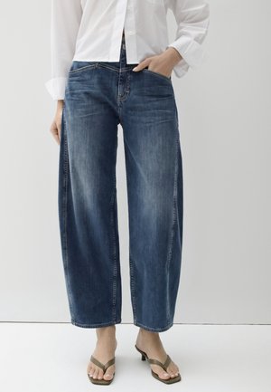 Persona che indossa jeans blu a gamba larga, una camicia bianca a maniche lunghe e sandali aperti con tacco basso, in piedi su uno sfondo semplice.